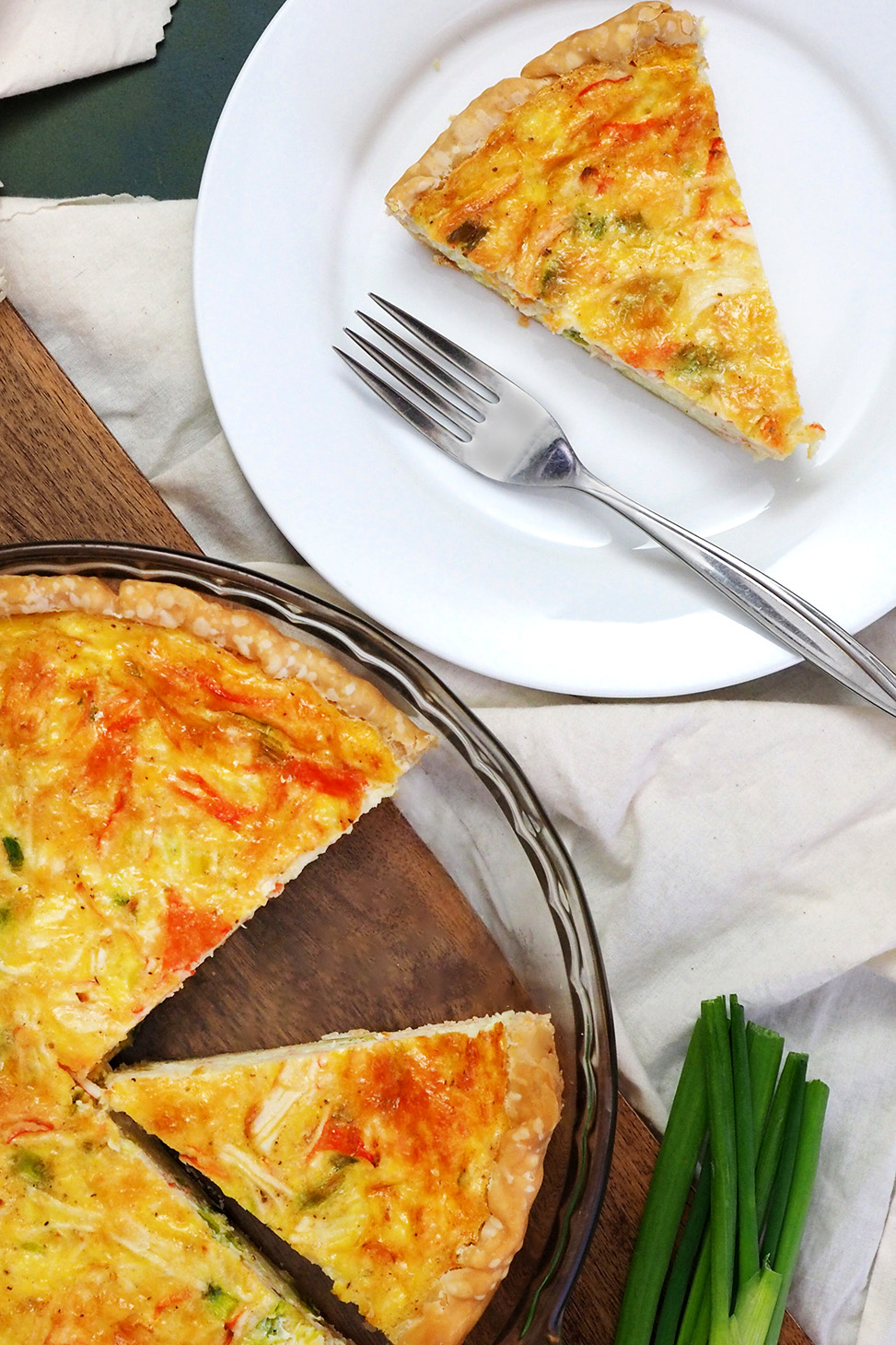 A white plate with a slice of crab quiche and a fork on it next to a pie plate full of crab quiche.