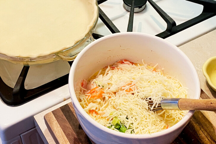 A bowl full of quiche ingredients with a whisk sticking out of it next to an empty, raw pie crust in a pie plate.