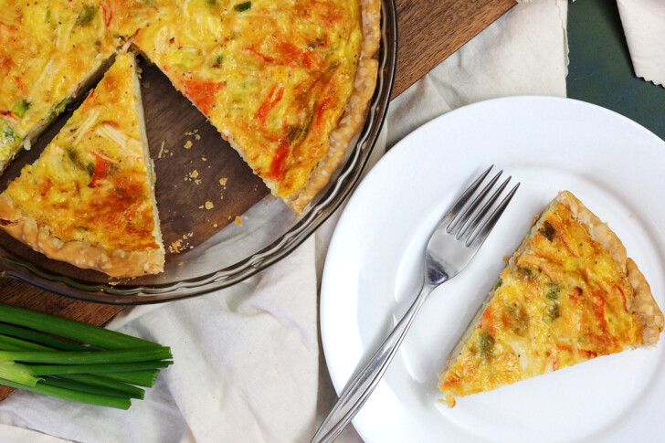 A slice of crab quiche on a white plate with a fork next to a glass pie plate with baked quiche in it.