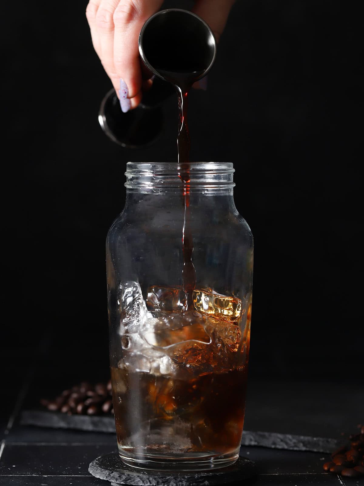 A hand pouring cold brew coffee from a jigger into a cocktail shaker filled with ice.