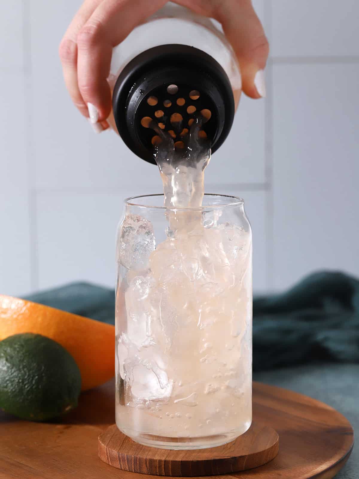 A hand pouring Paloma cocktail into a glass filled with ice. 