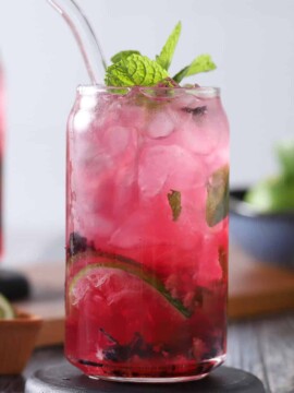 A glass filled with blueberry mojito cocktail, fresh limes, fresh mint, and crushed blueberries.