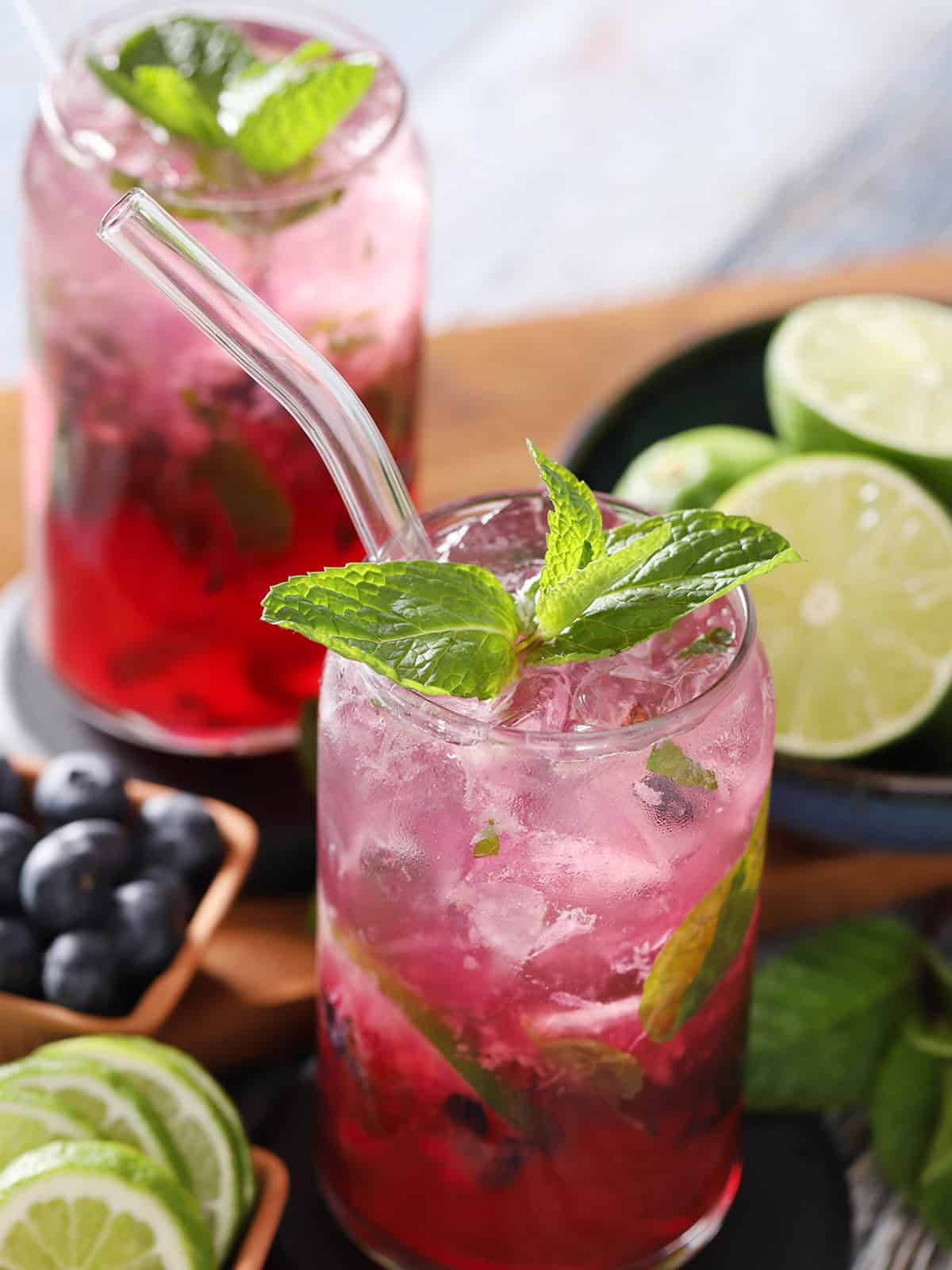 Glasses filled with blueberry mojito cocktail, fresh limes, fresh mint, and crushed blueberries.