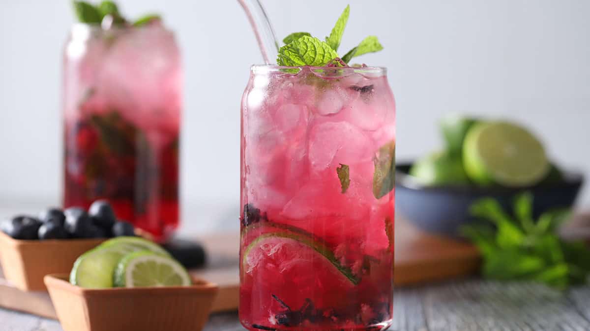 Glasses filled with blueberry mojito cocktail, fresh limes, fresh mint, and crushed blueberries.