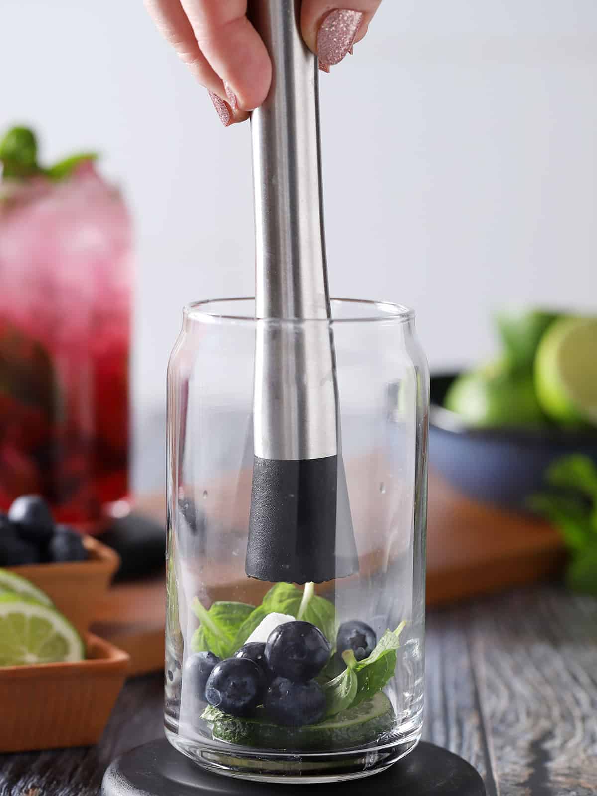 A hand placing a muddler into a glass with blueberries, fresh mint, lime slices and a sugar cube in the bottom. 