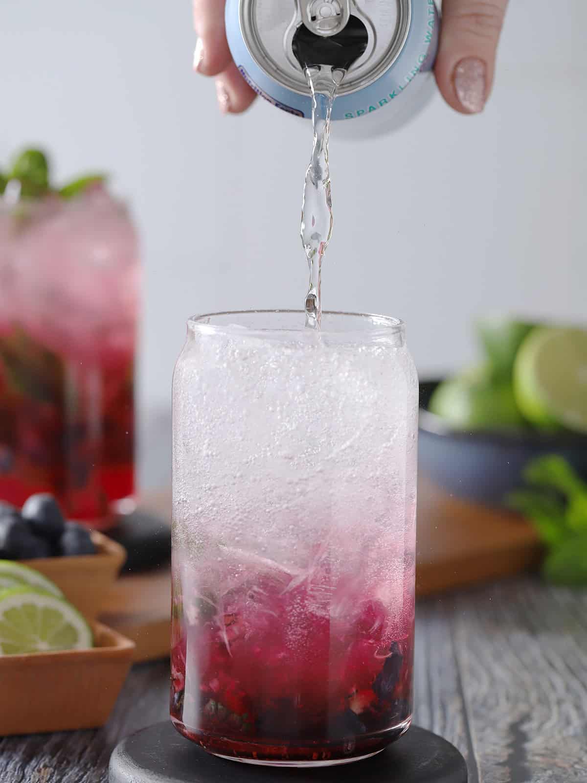 A hand pouring sparkling water from a can into a glass filled with blueberry syrup, rum, and ice. 