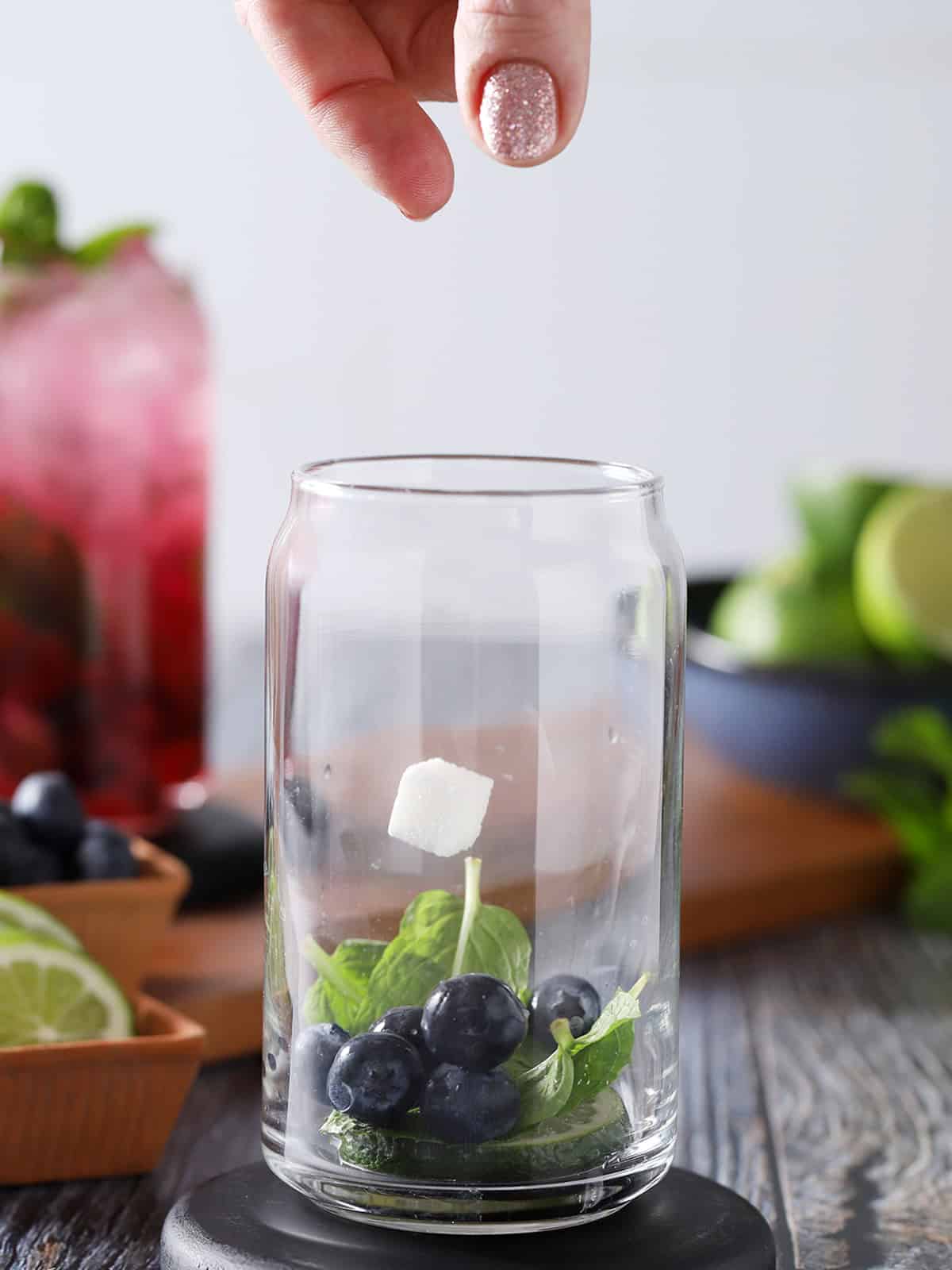 A sugar cube being dropped into a glass filled with fresh mint, blueberries, and lime slices. 