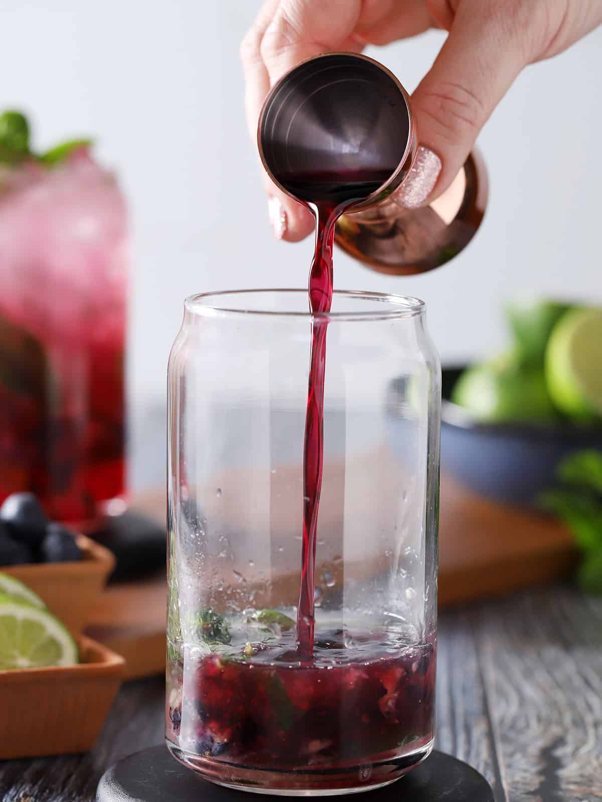 A hand pouring blueberry syrup from a jigger into a glass. 