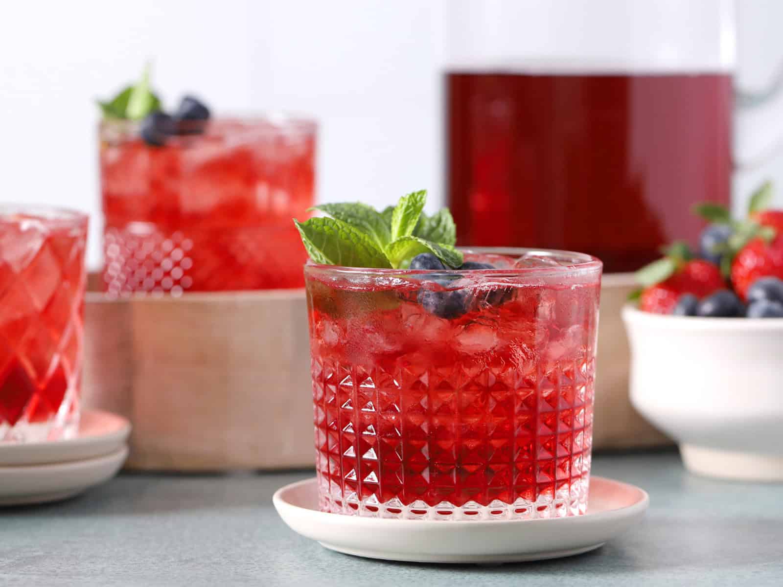 Glasses filled with red drinks and fruit, with a pitcher of red drinks in the background.