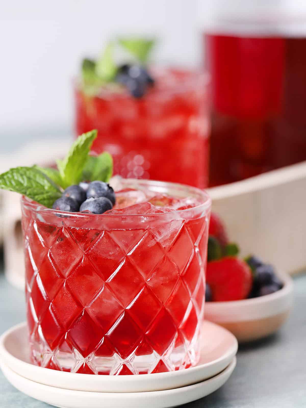 Glasses filled with red drinks and fruit, with a pitcher of red drinks in the background.