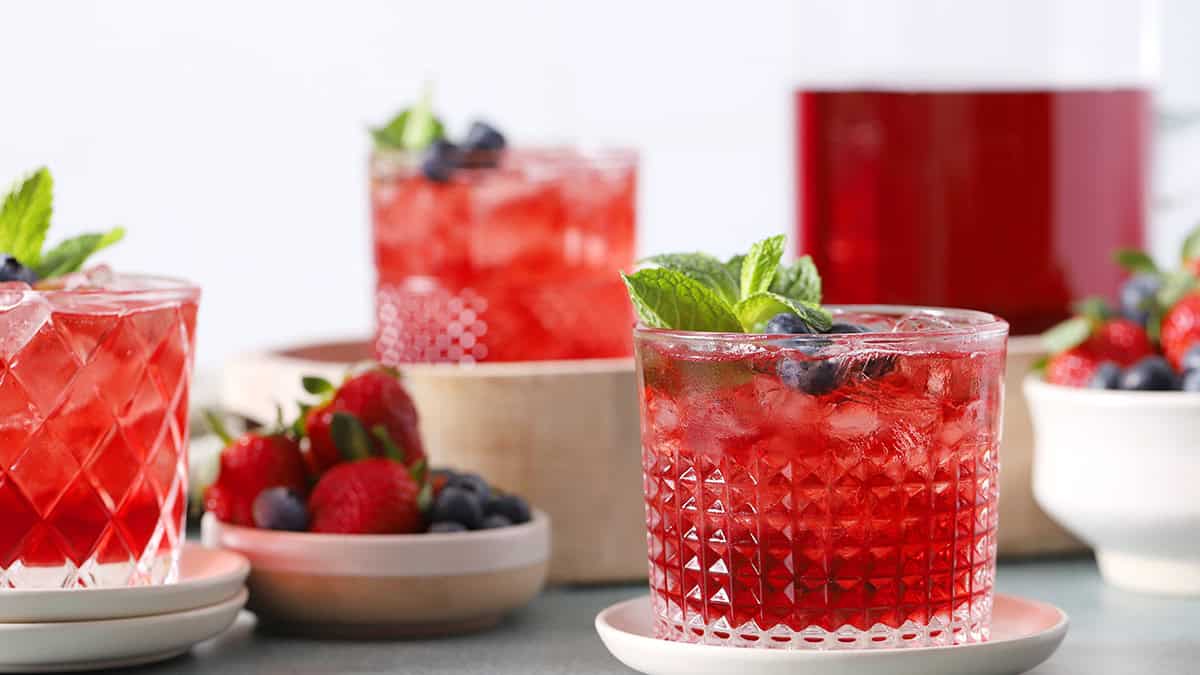 Glasses filled with red drinks and fruit, with a pitcher of red drinks in the background.
