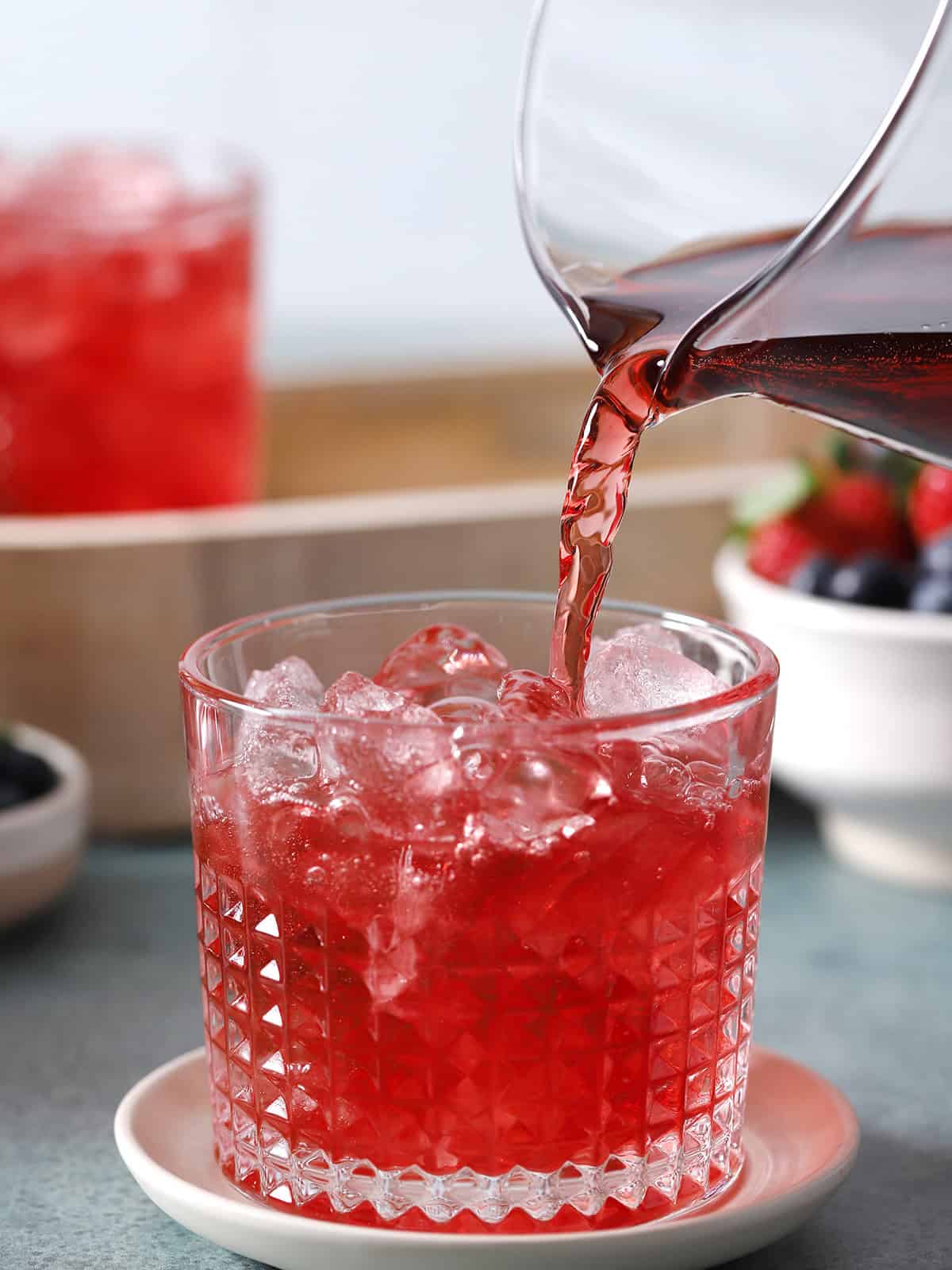A pitcher of red mocktail pouring into a glass filled with ice. 