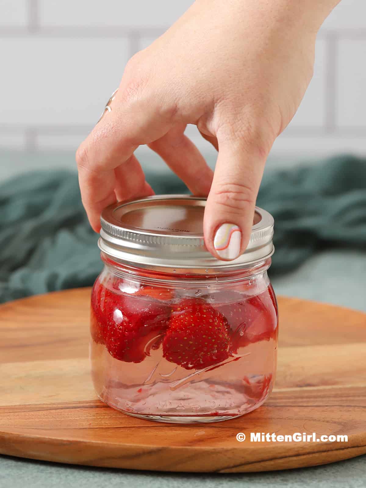 A hand screwing the lid onto a jar filled with vodka and strawberries.