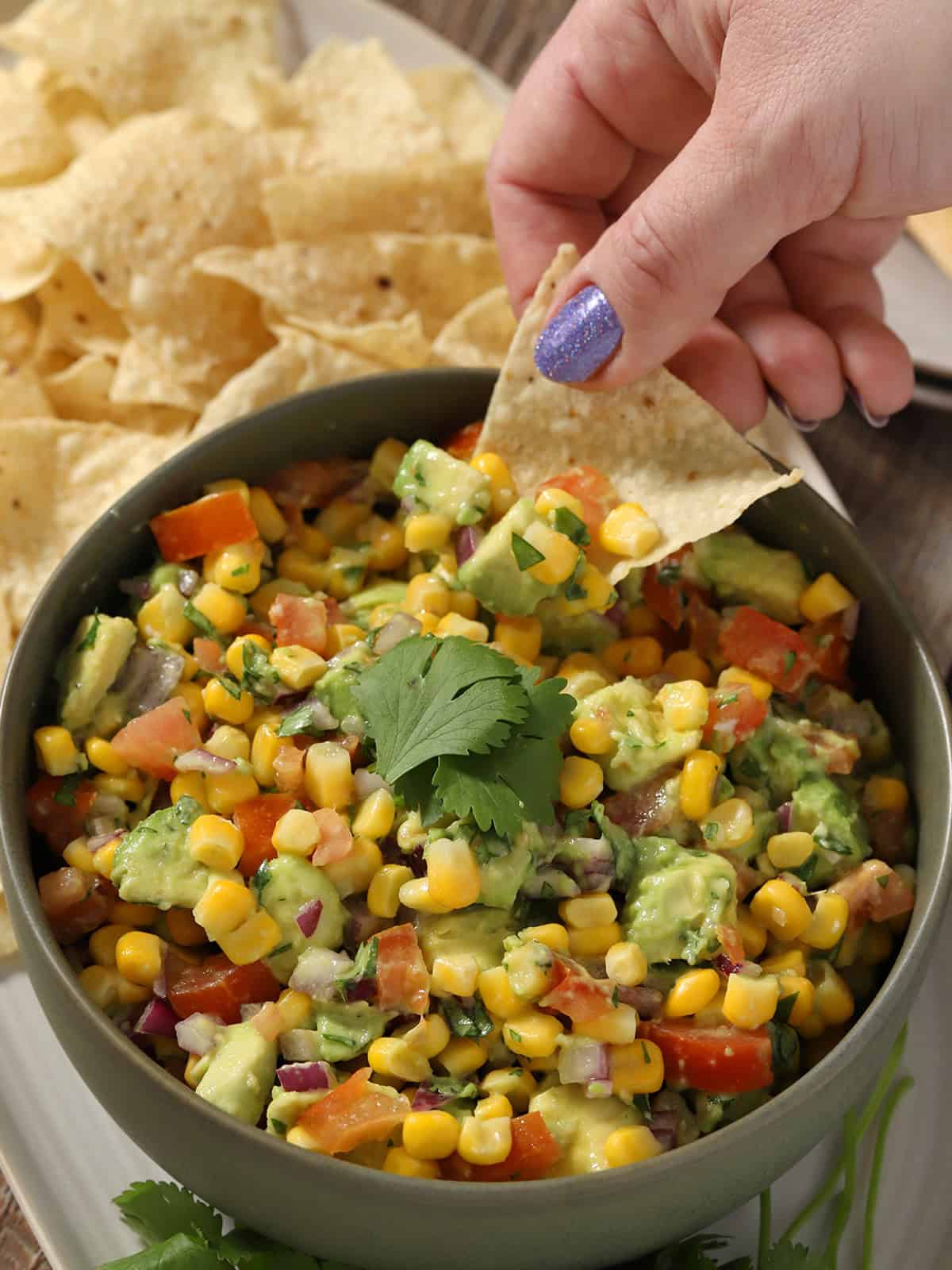 A hand scooping a tortilla chip into a bowl of corn avocado salsa.