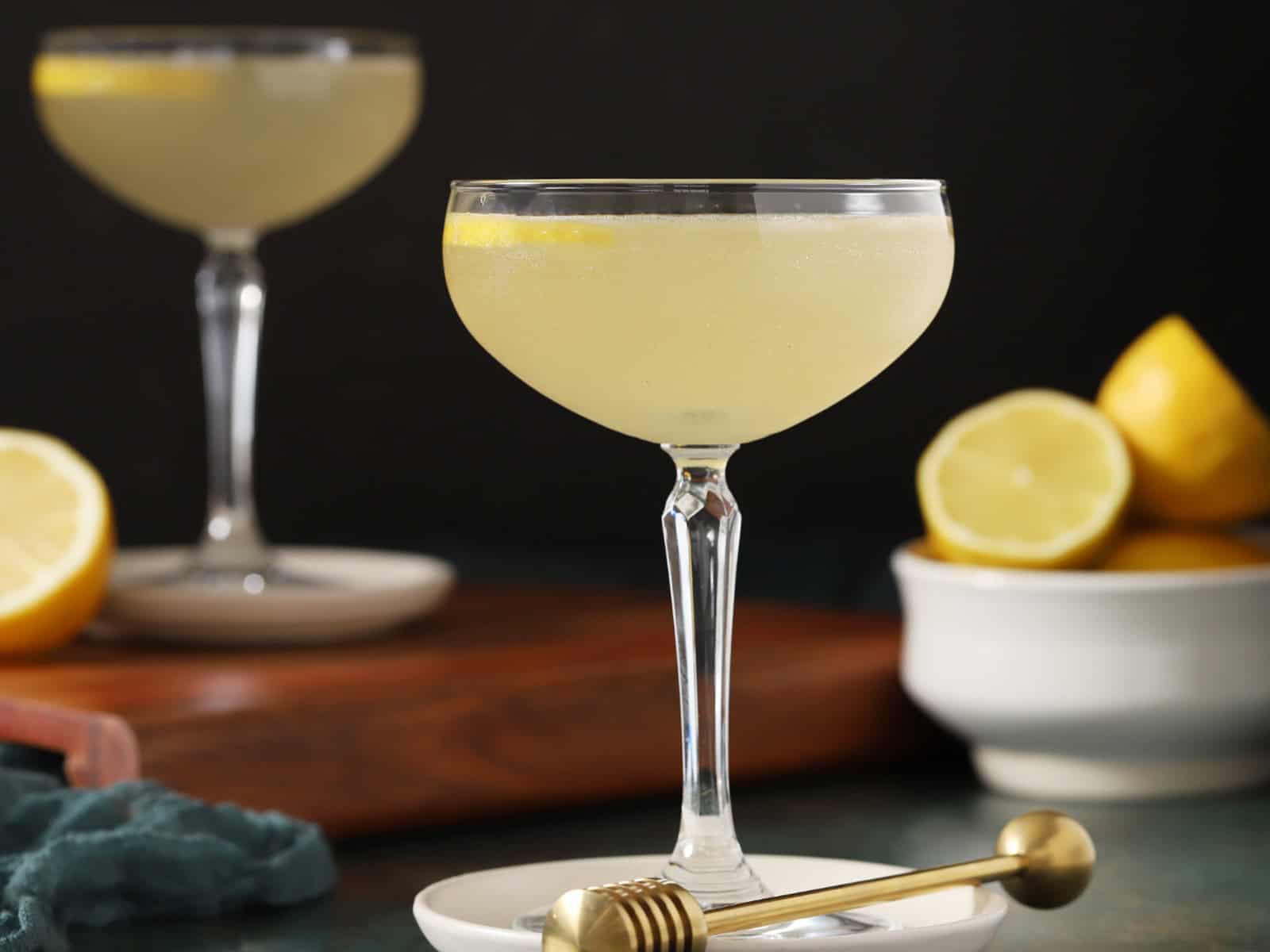 A pair of coupe glasses filled with bee's knees cocktail with fresh lemons.