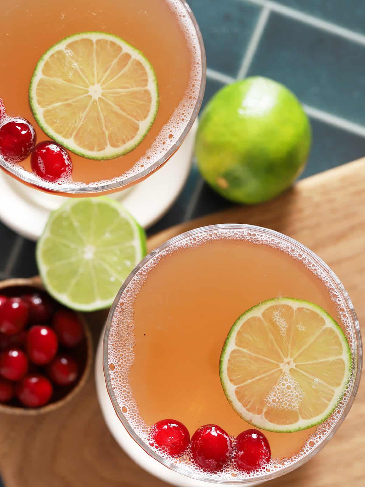 Looking down at two cocktail glasses filled with cosmos, garnished with lime slices and cranberries. 