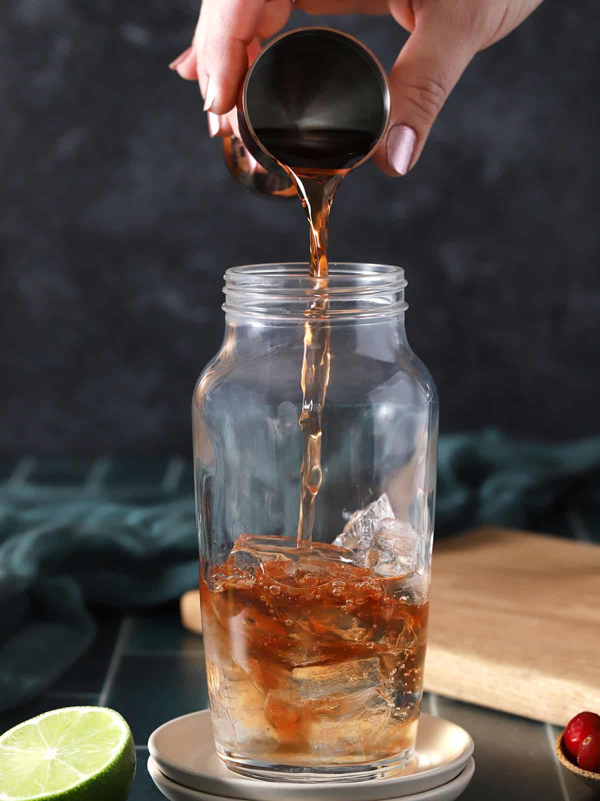 A hand pouring cranberry juice from a jigger into a cocktail shaker with ice. 
