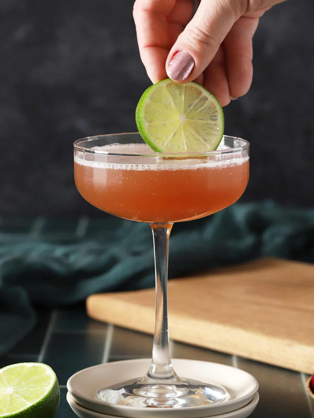 A hand placing a lime wheel garnish into a coupe glass filled with cosmopolitan cocktail.