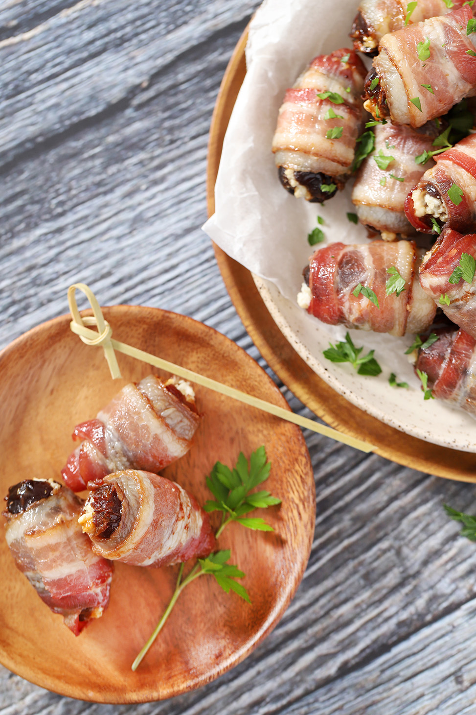 A wooden plate of 2 bacon-wrapped dates next to a larger platter of more bacon wrapped dates.
