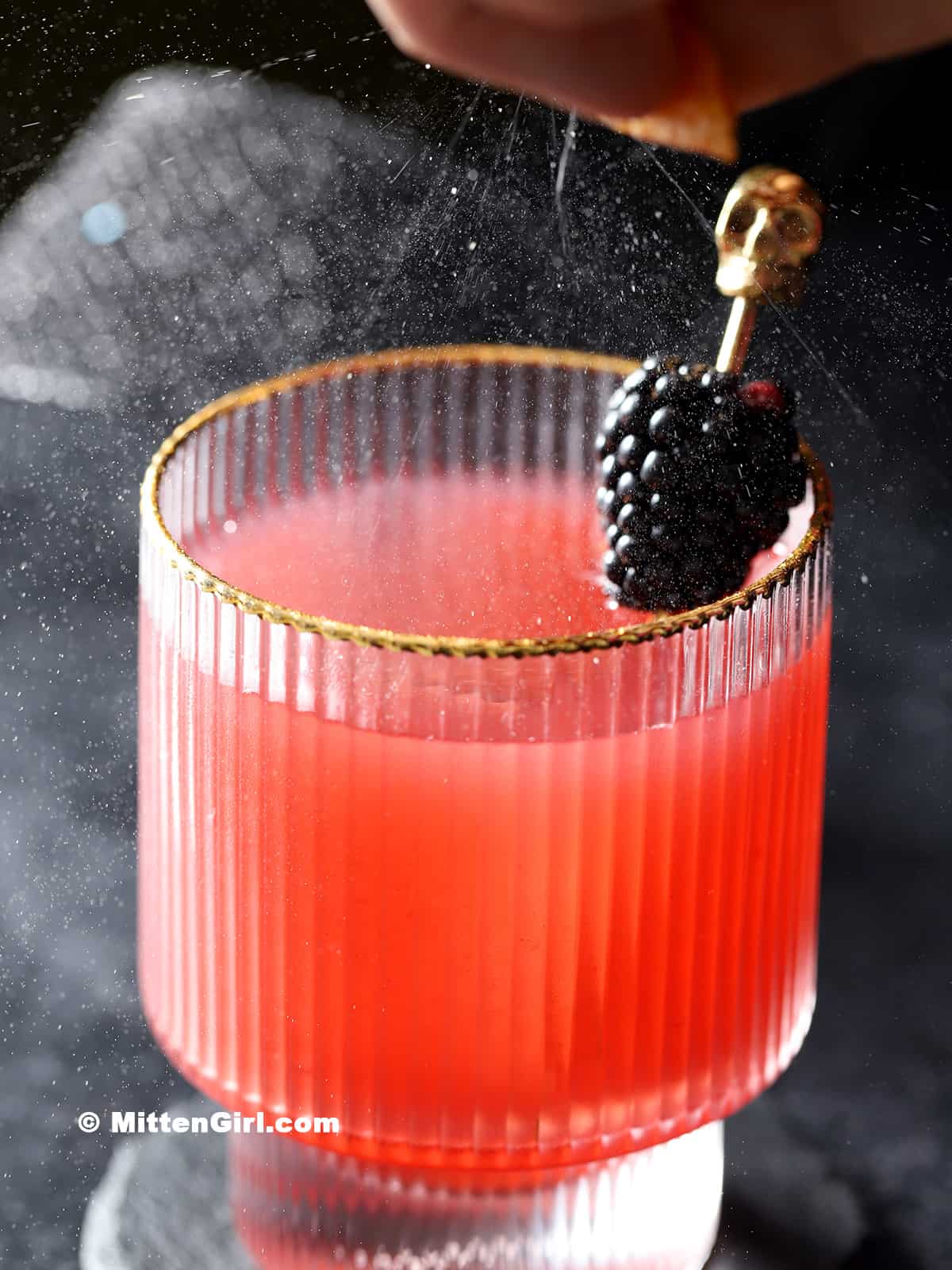 Orange oil from a peel being expressed over a glass of blackberry bourbon cocktail.