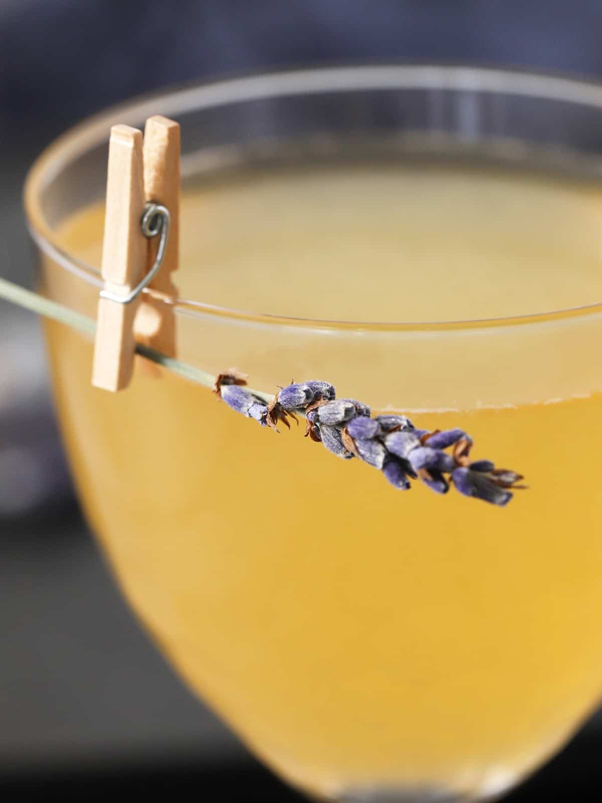A glass of cocktail garnished with a small sprig of lavender flowers held on by a tiny clothespin.
