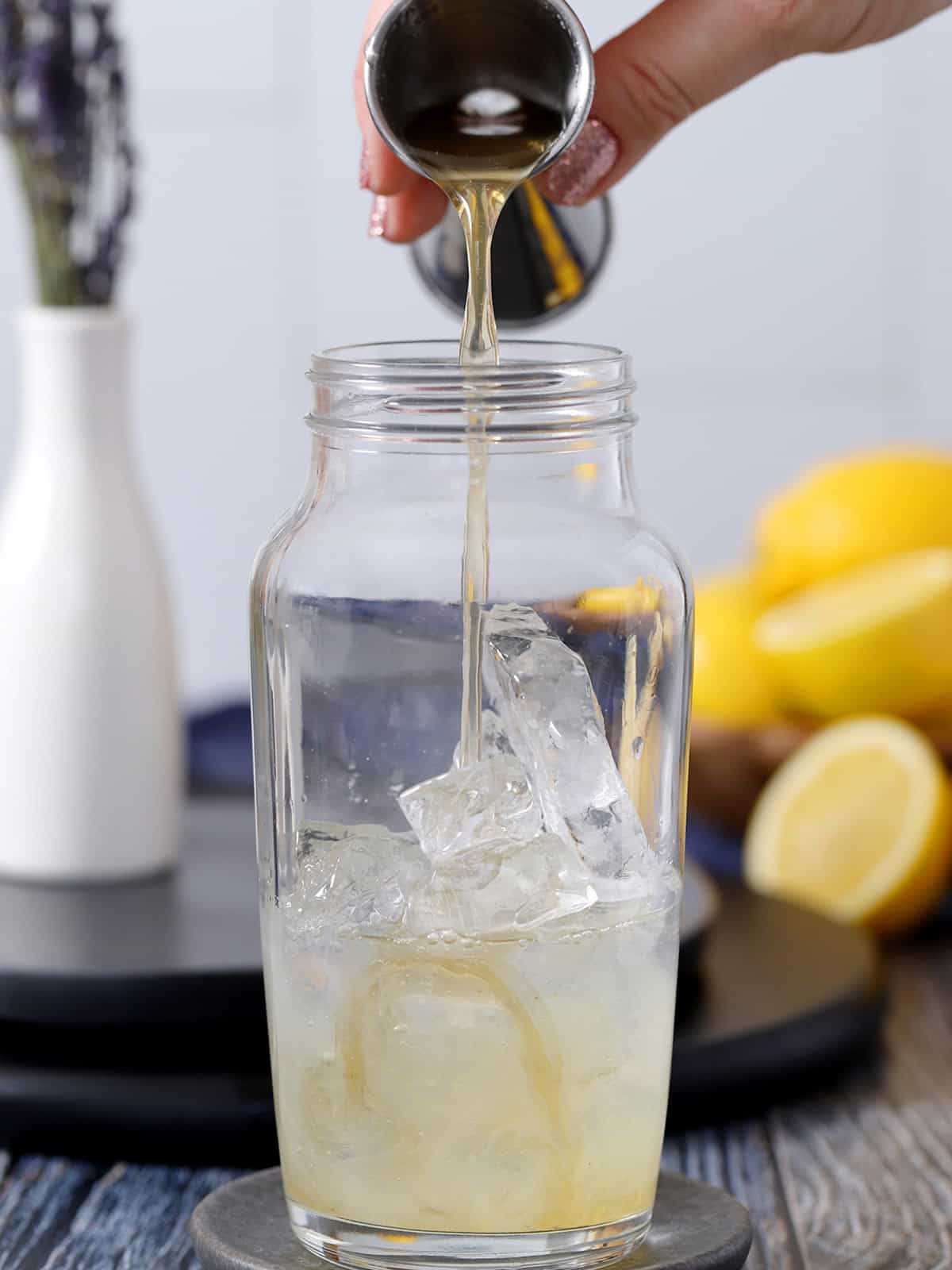 A hand pouring honey syrup into a cocktail shaker filled with gin and ice.