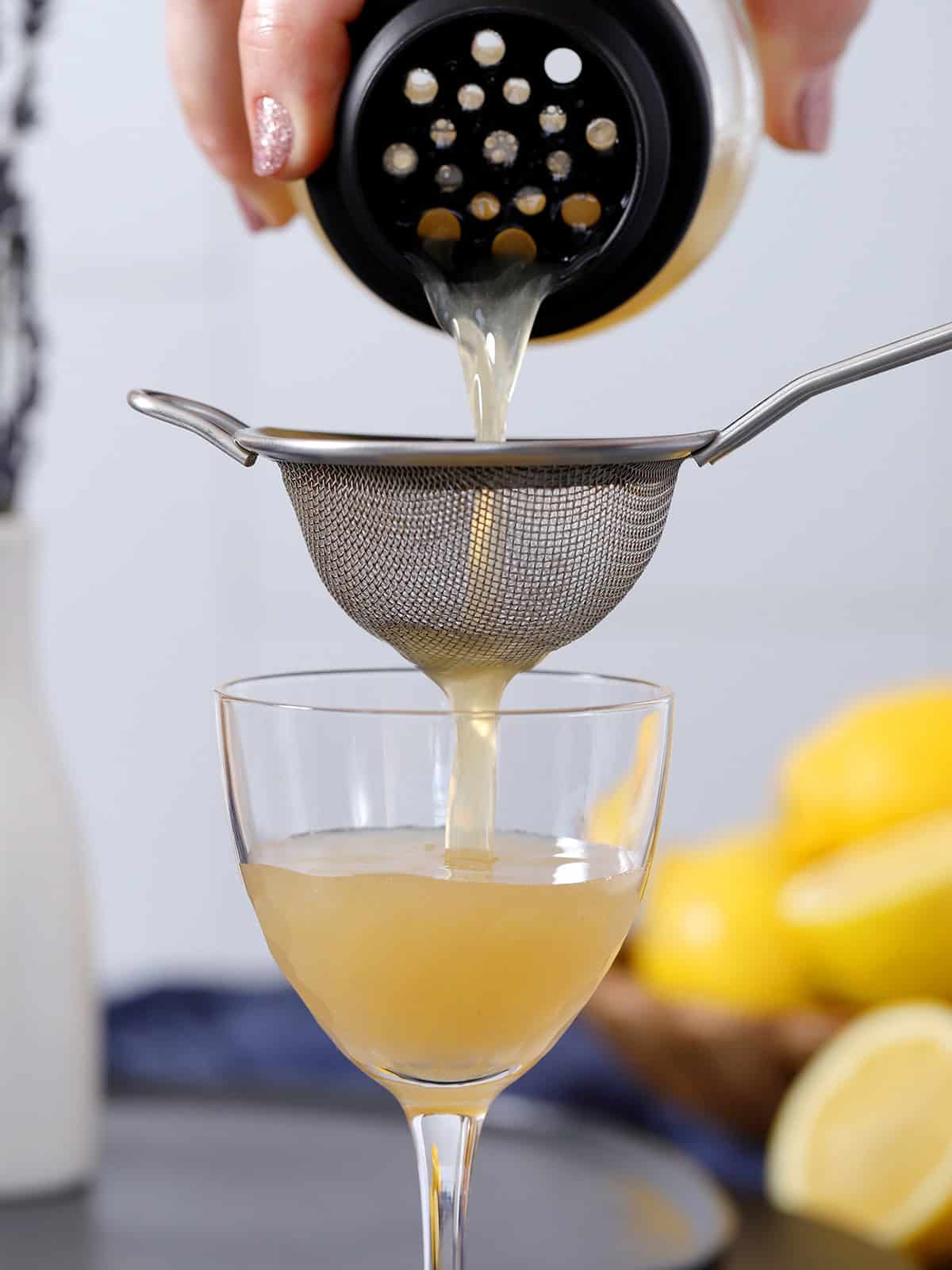 A hand pouring a lavender bee's knees cocktail from a shaker through a fine mesh strainer and into a Nick and Nora glass.