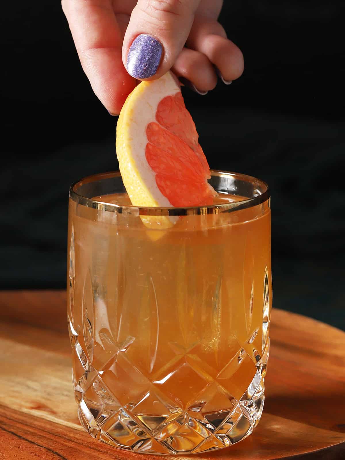 A hand placing a slice of grapefruit into a glass of bourbon cocktail. 