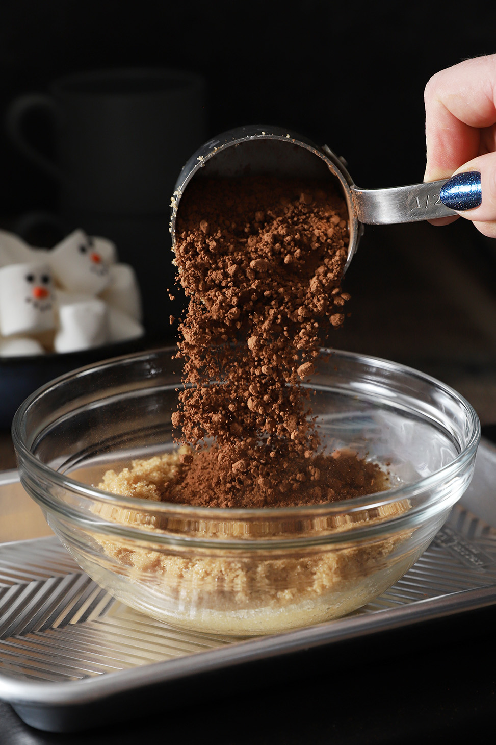 A hand pouring cocoa powder into a bowl filled with sugars.