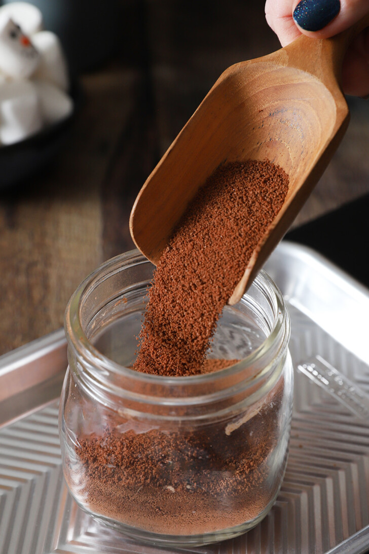 Hot chocolate mix being poured into a glass jar using a wooden scoop.