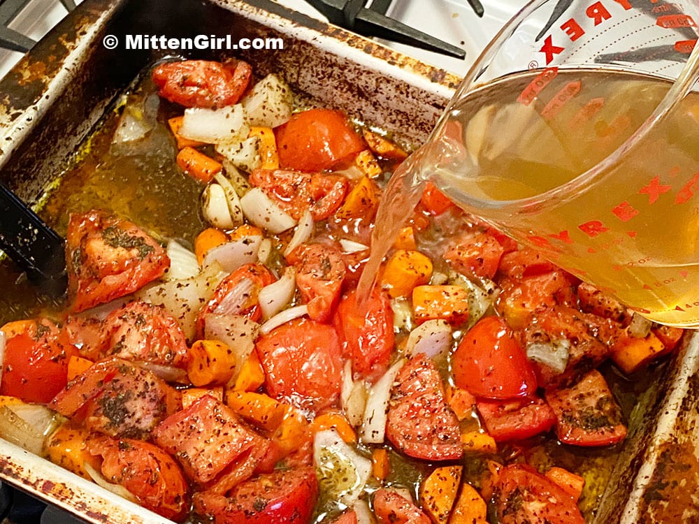 Pouring chicken broth in a pan of roasted tomatoes and other vegetables