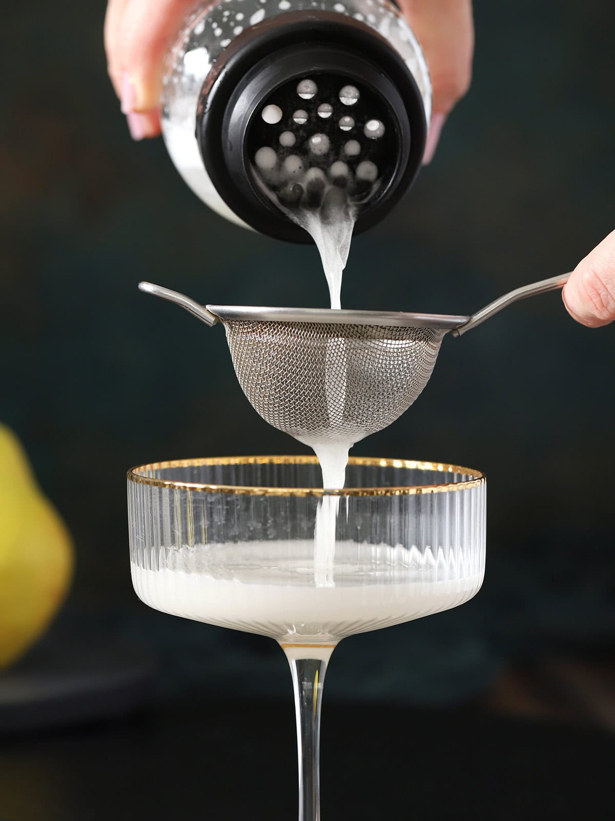 A hand pouring pear sour from a cocktail shaker through a fine mesh strainer into a coupe glass below.