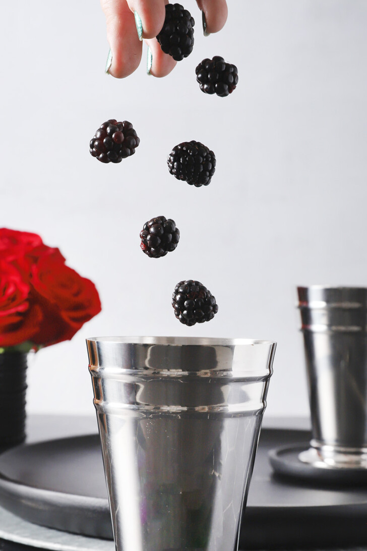 A hand dropping fresh blackberries into a silver julep cup.