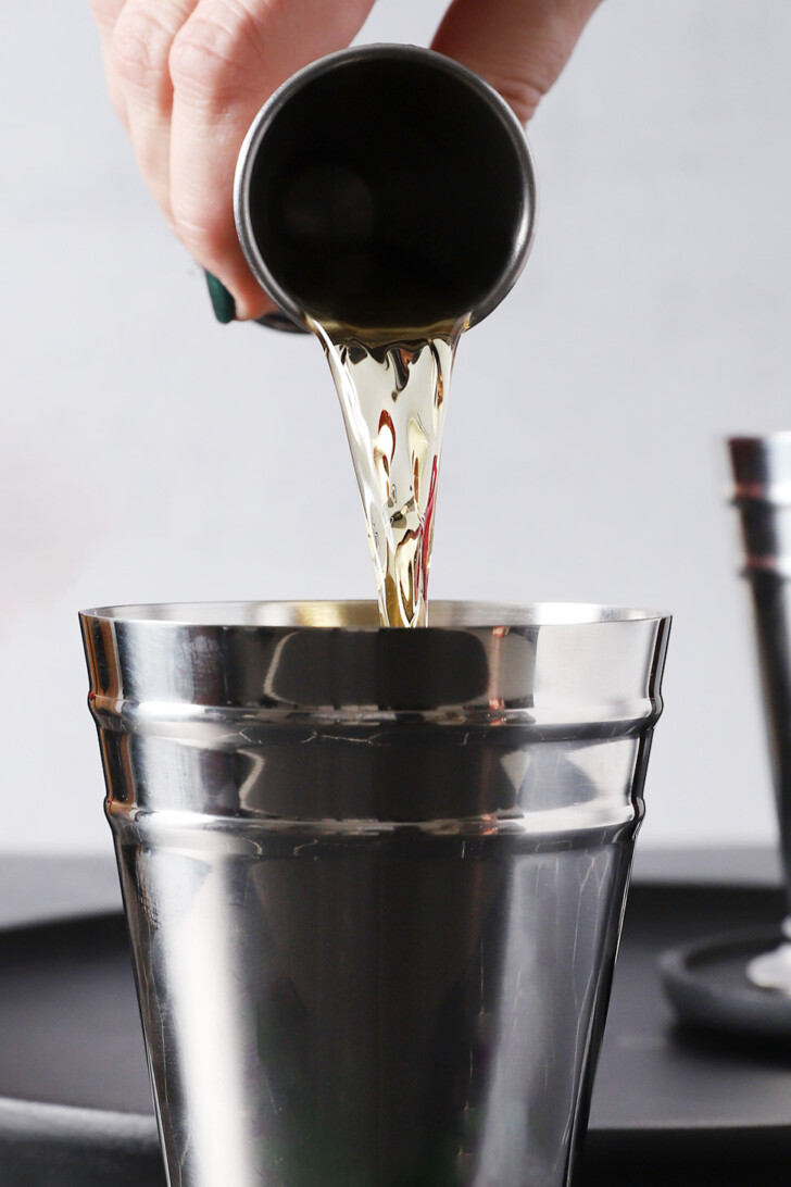 A hand pouring bourbon from a silver jigger into a silver julep cup.