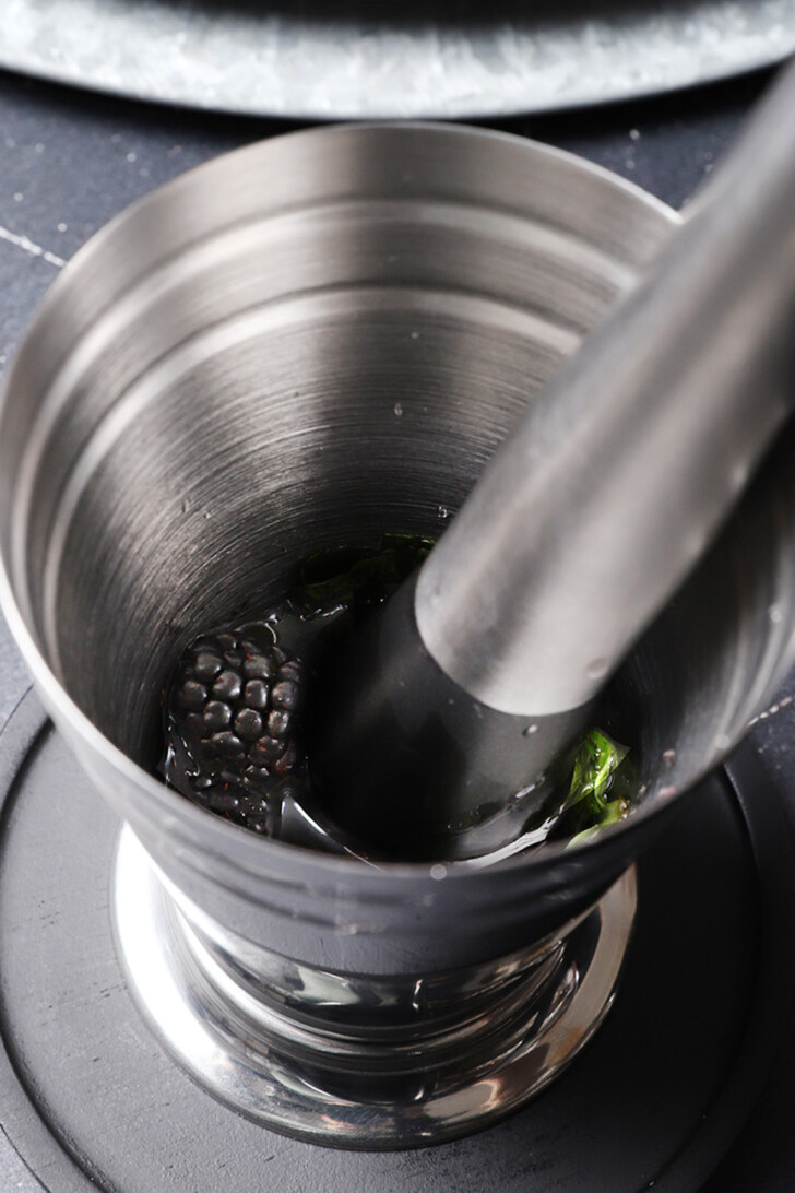 A muddler in a julep cup with blackberries and mint leaves.