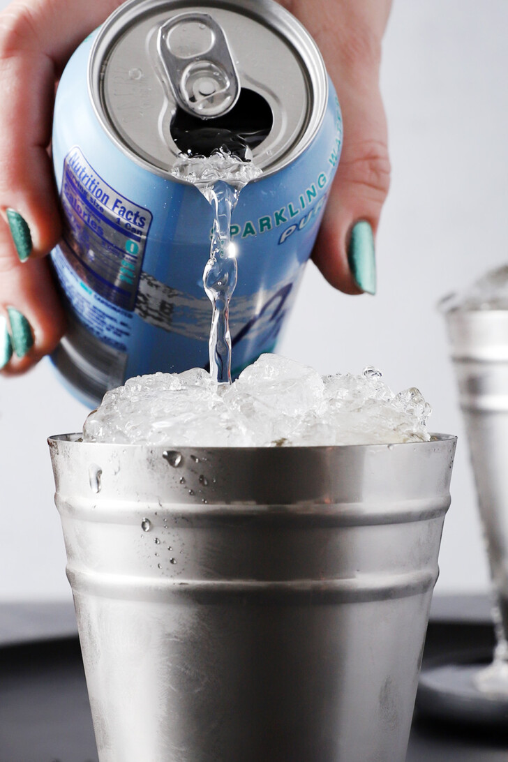 A hand pouring soda water into a julep cup heaped with ice.