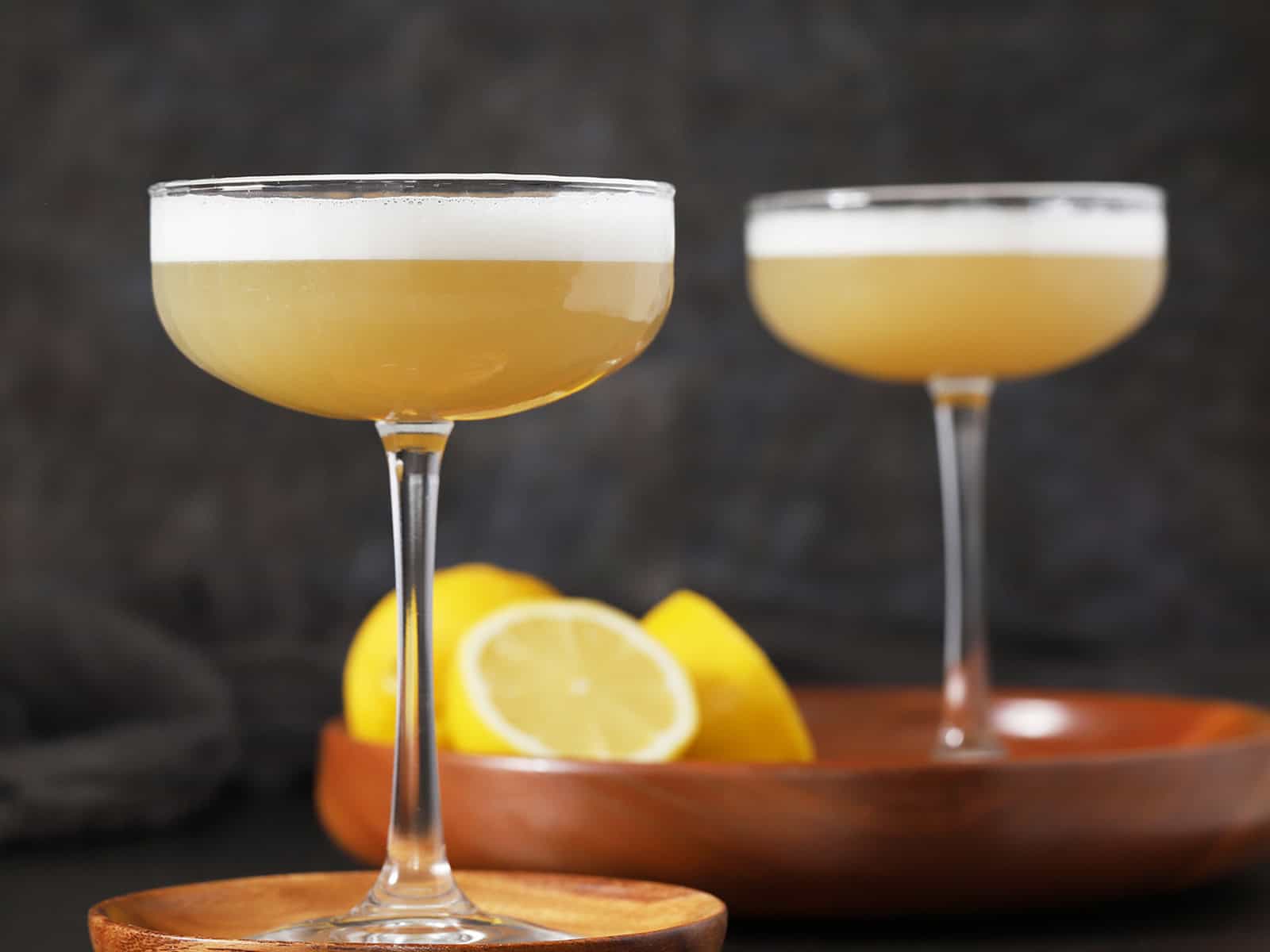 Two glasses of ginger bourbon sour cocktail on wooden trays with lemons.