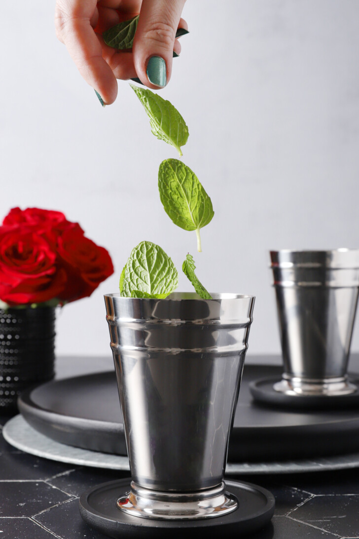 A hand dropping fresh mint leaves into a silver julep cup.
