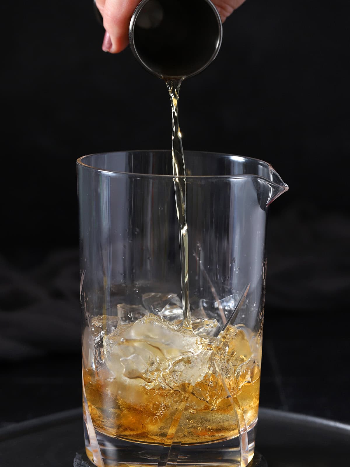 A hand pouring bourbon from a jigger into a mixing glass with ice.