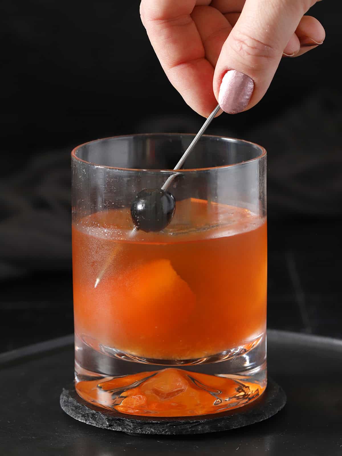 A hand lowering a cocktail pick with a cherry on it into a rocks glass filled with old fashioned cocktail.