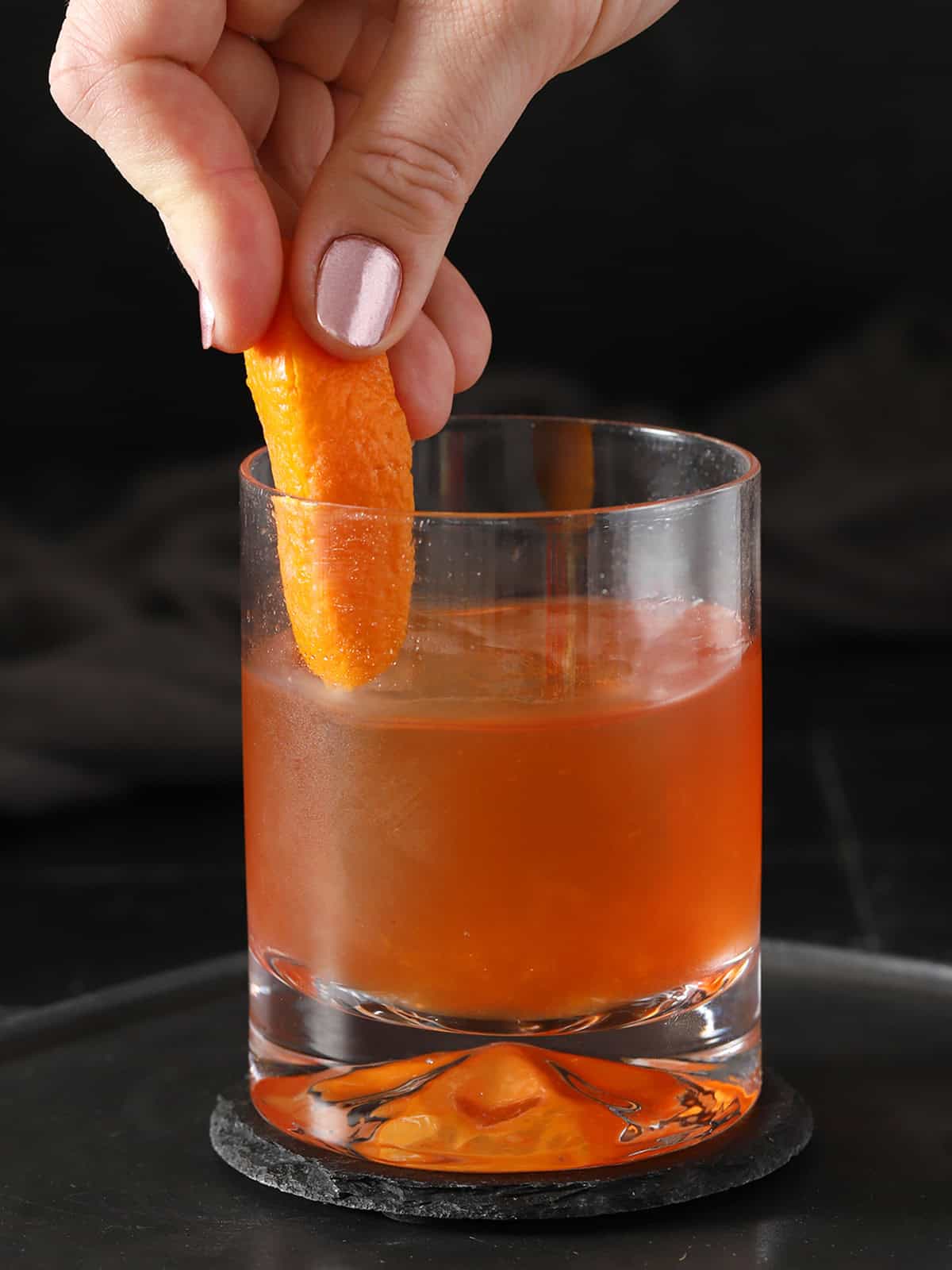 A hand running slice of orange peel around the inside of a rocks glass filled with old fashioned.