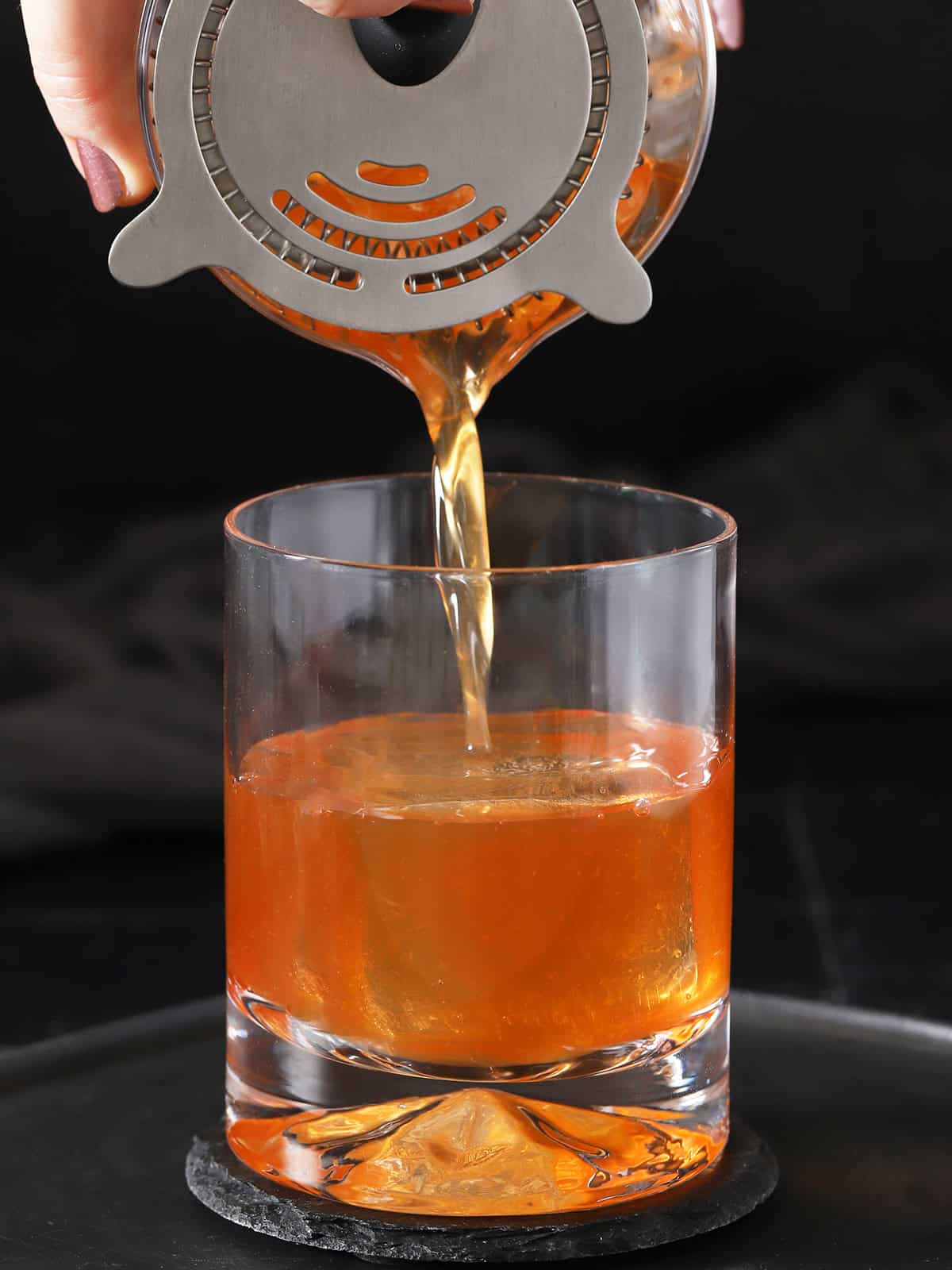 A hand holding a hawthorne stainer over a mixing glass and pouring old fashioned cocktail into a rocks glass.