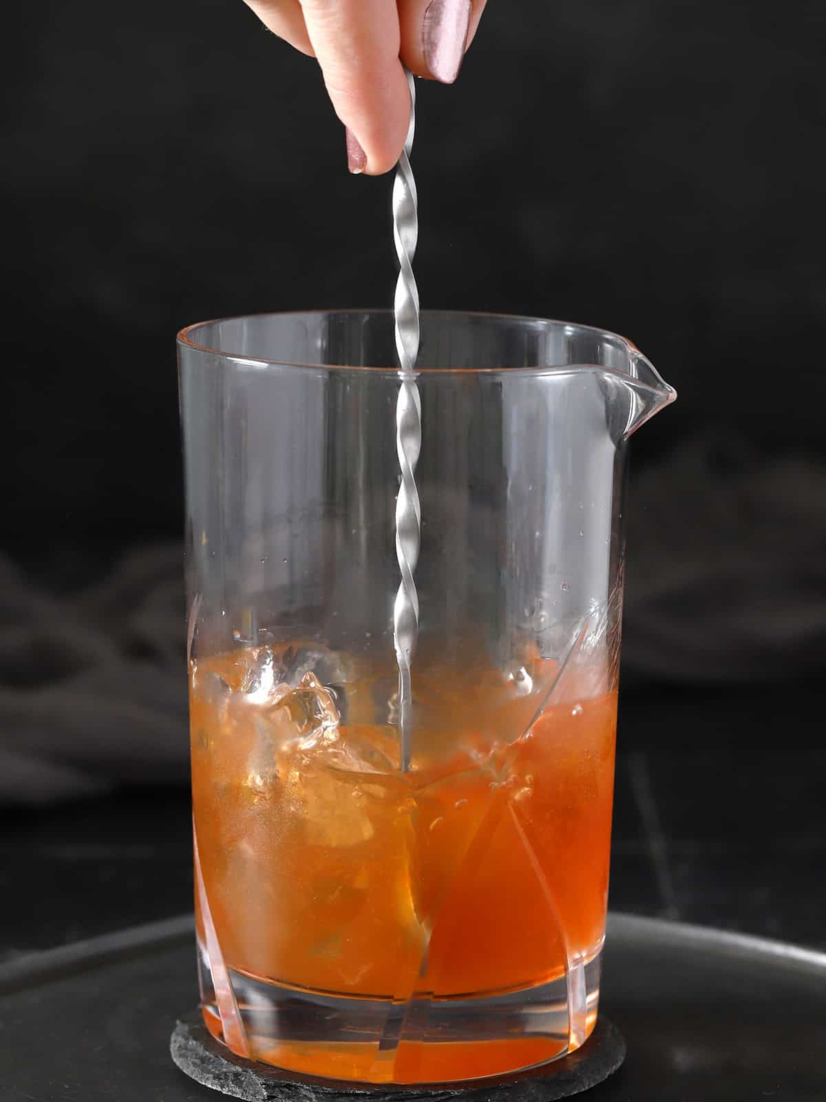 A hand using a bar spoon to stir cocktail in a mixing glass.
