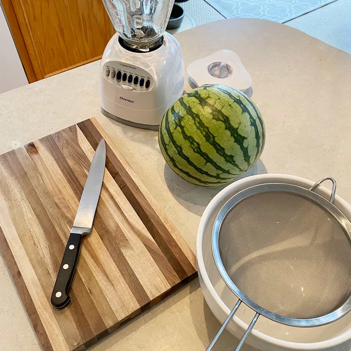 A blender, watermelon, knife, cutting board, bowl, and strainer sitting on the counter.