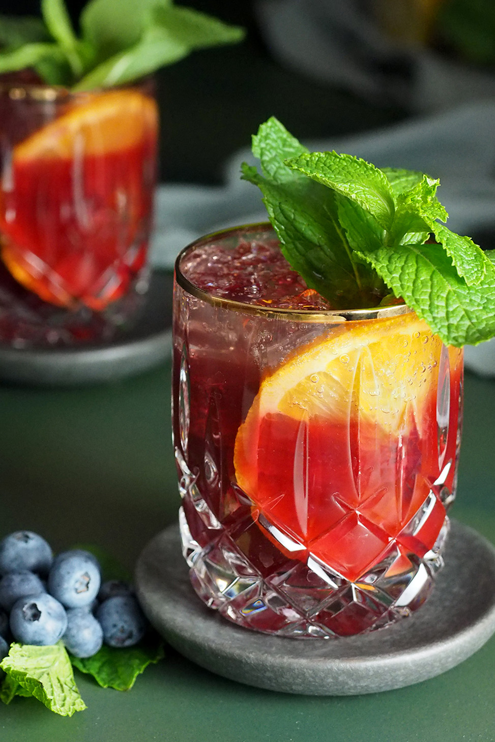 A rocks glass filled with blueberry citrus julep cocktail garnished with a large slice of orange and large sprig of mint.