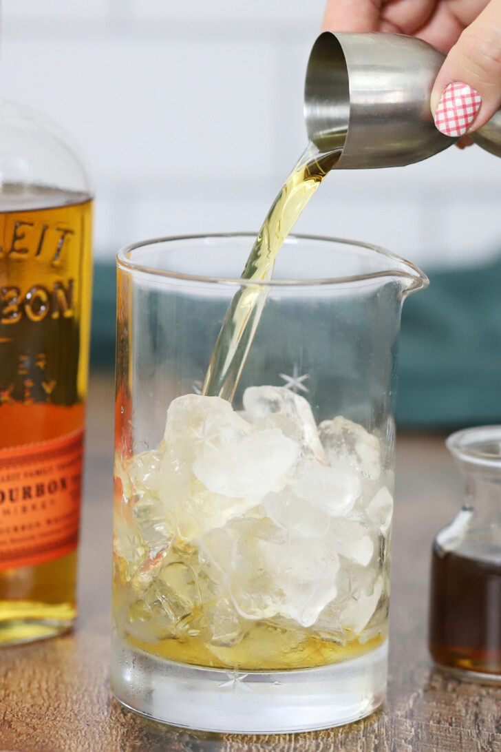 A hand pouring bourbon from a jigger into a mixing glass.