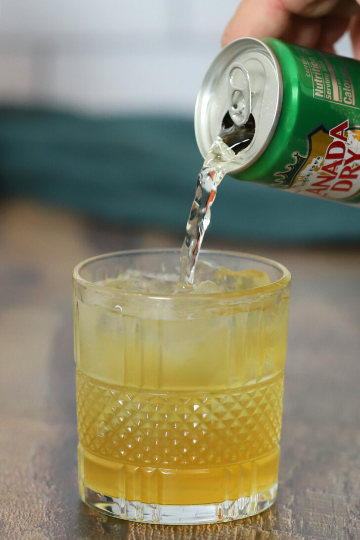 A hand pouring ginger ale from a small can into a rocks glass filled with bourbon ginger cocktail and ice.