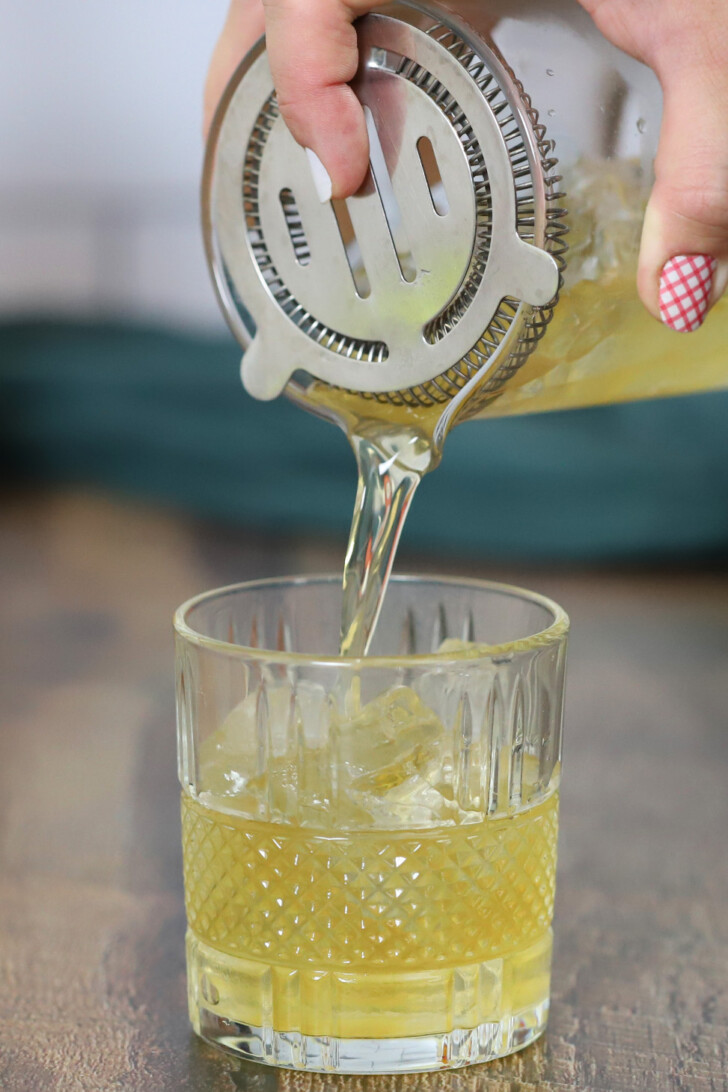 A hand pouring a bourbon ginger cocktail into a rocks glass filled with ice.