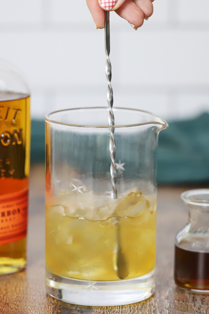 A hand stirring a bourbon ginger cocktail in a mixing glass with ice.