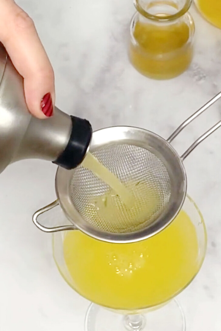 A hand pouring a honey orange gin cocktail from a cocktail shaker into a coupe glass.